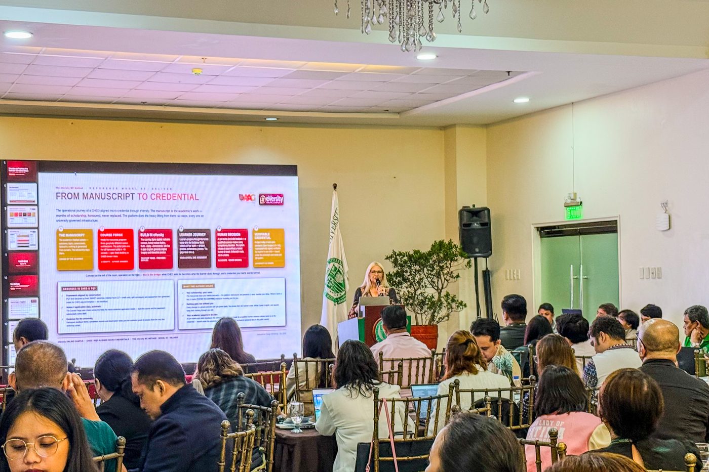 Dr. Ingrid Ursua presenting at the front of the room beside a slide titled From Manuscript to Credential